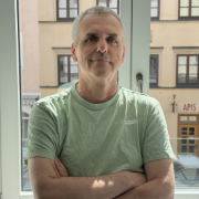 Člen týmu Dlubal stojící před kancelářskými okny se zkříženýma rukama a přátelským výrazem.