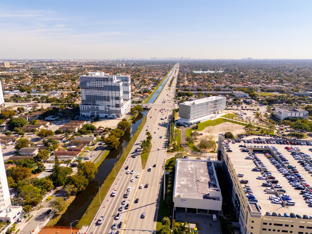 Blick auf eine belebte Straße vor dem Campus der FIU in Miami mit Palmen und vorbeiziehenden Autos im Vordergrund.