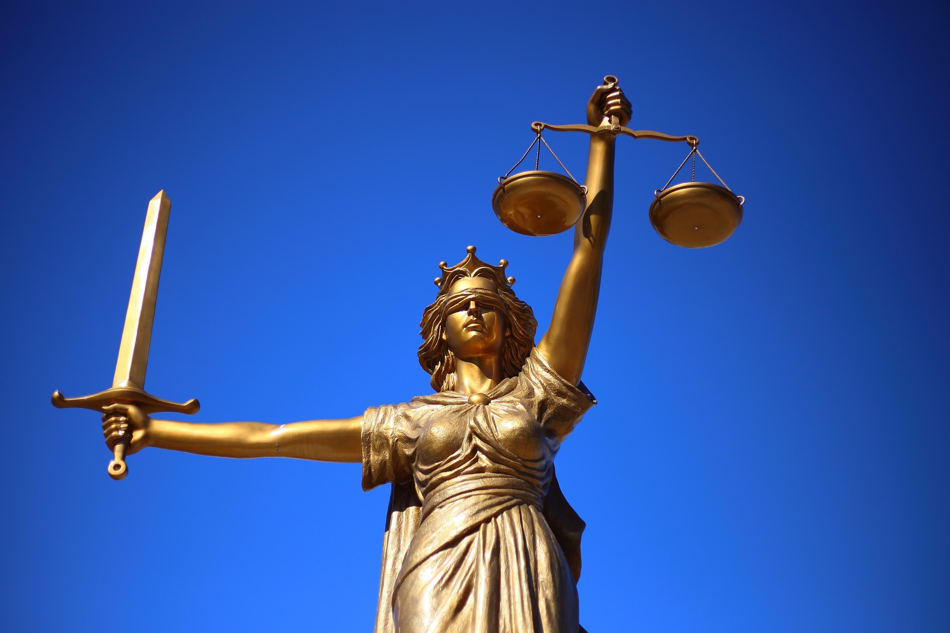 Goldene Justitia-Statue mit Waage und Schwert vor blauem Himmel als Symbol für die komplexe Rechtsprechung und Gesetzesdurchsetzung im Bauwesen.
