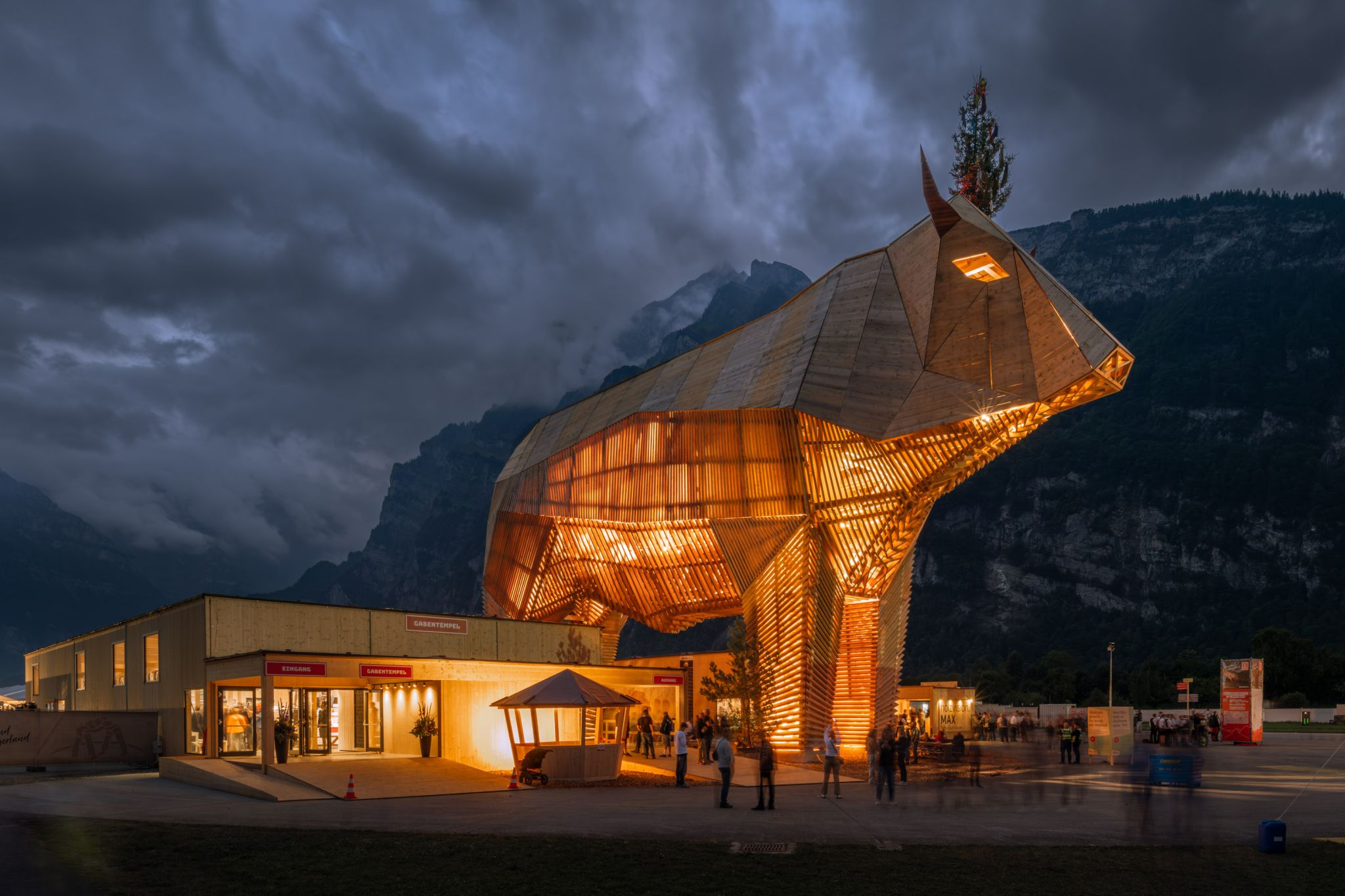 The illuminated wooden bull “Muni MAX” in Switzerland, a pagoda-like timber structure with a stained timber facade set against an alpine mountain backdrop at dusk.
