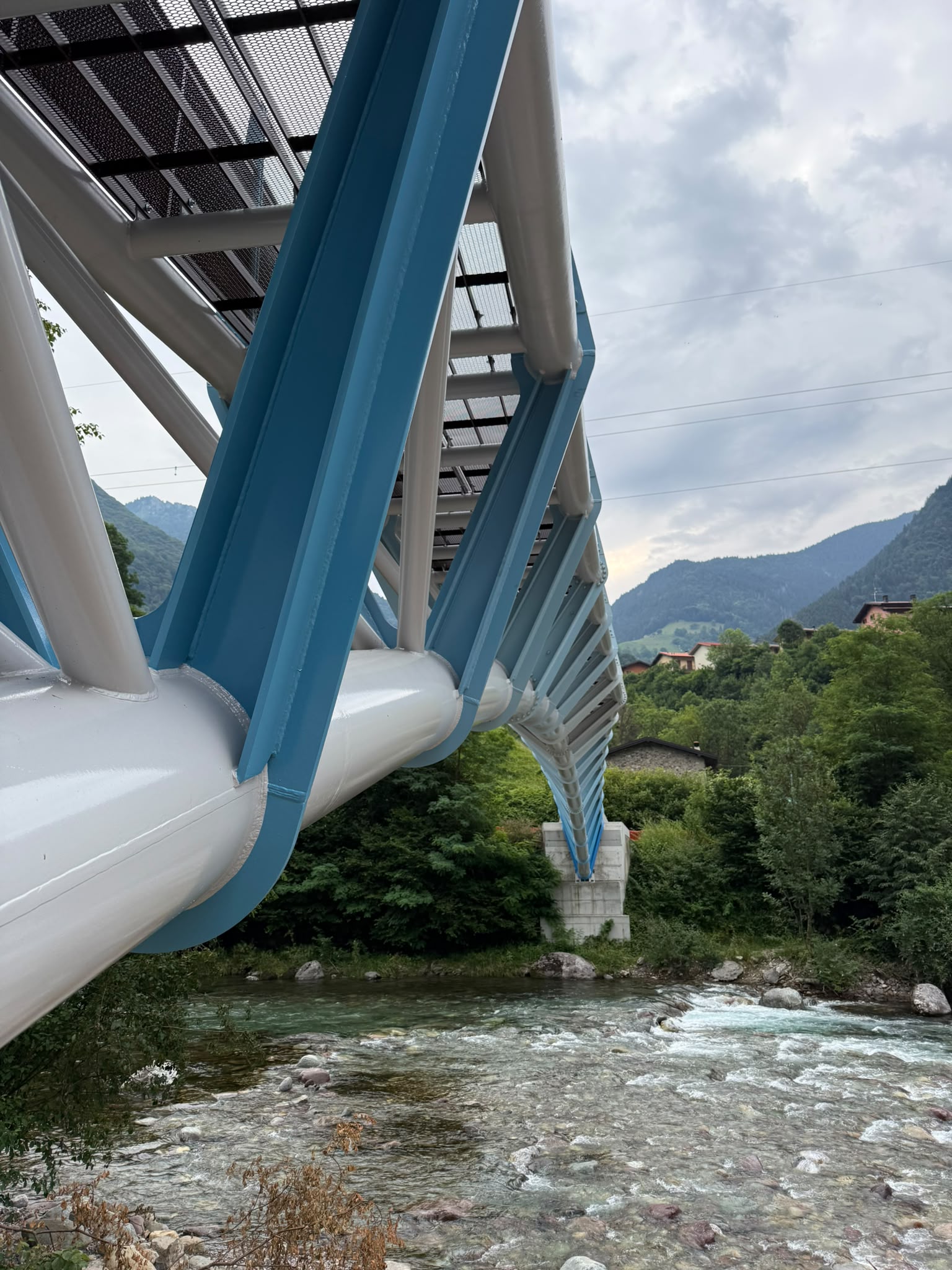 Steel structure of a curved pedestrian bridge over a river, with an alpine landscape in the background