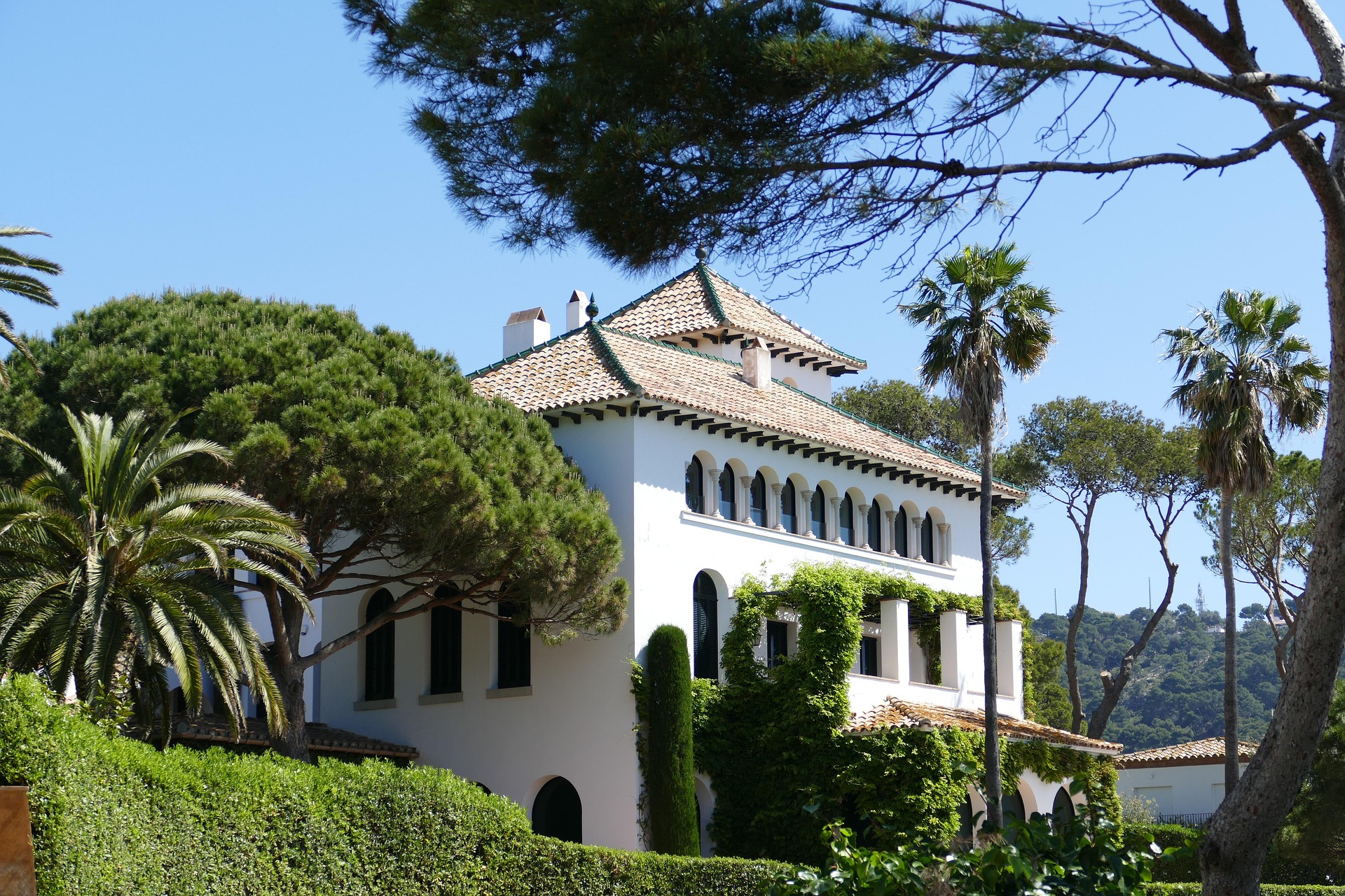 Villa méditerranéenne à façade blanche, toit en tuiles et fenêtres en arcades, entourée de pins et de palmiers dans le paysage ensoleillé de la Costa Brava.