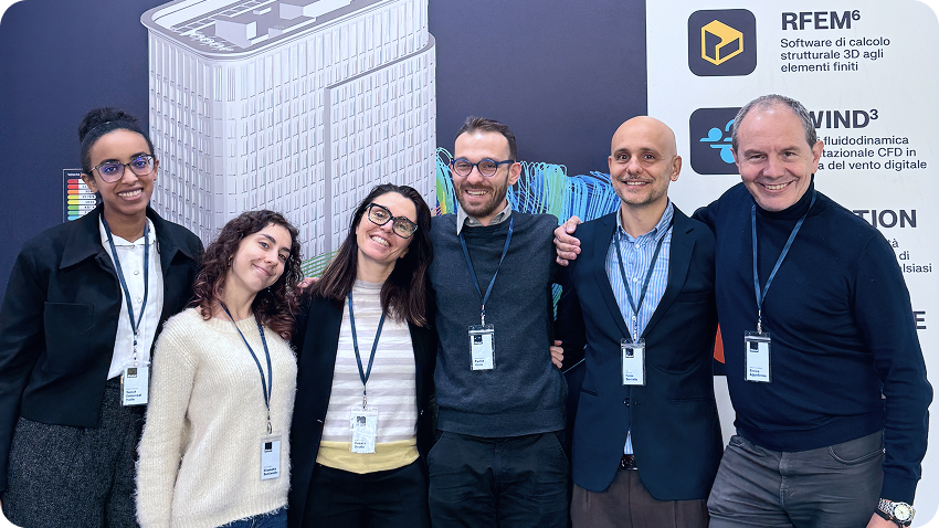 Colleghi italiani riuniti presso lo stand di una fiera durante un evento, discutendo concetti di ingegneria strutturale con i visitatori.