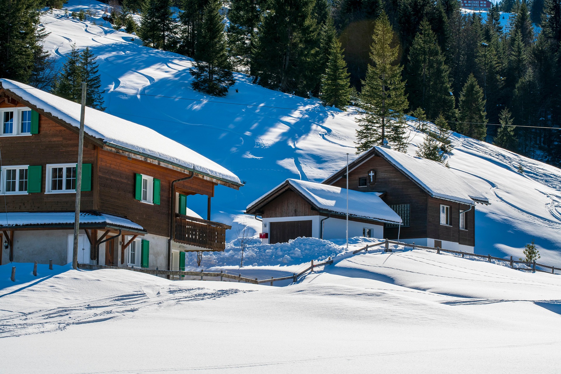 Due case tradizionali in legno con persiane verdi e coperture innevate in un paesaggio alpino invernale con tempo sereno e soleggiato.