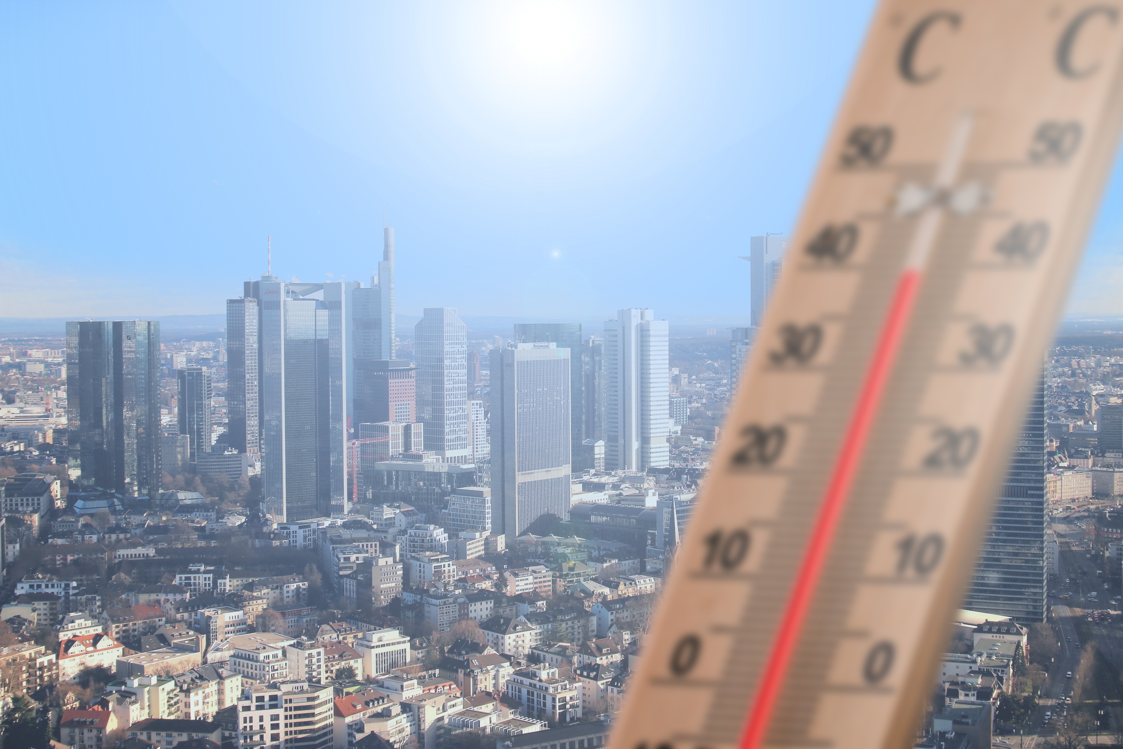 Captura de uma linha do horizonte de uma grande cidade com um termómetro em primeiro plano, indicando temperaturas elevadas. A imagem ilustra os efeitos das ondas de calor nas estruturas urbanas e a crescente importância da simulação térmica de edifícios.